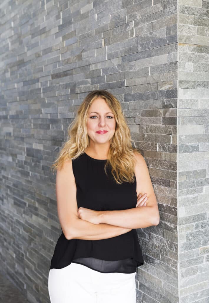 Kathryn Lott is seen leaning against a stone wall. She's wearing a black top and white pants.