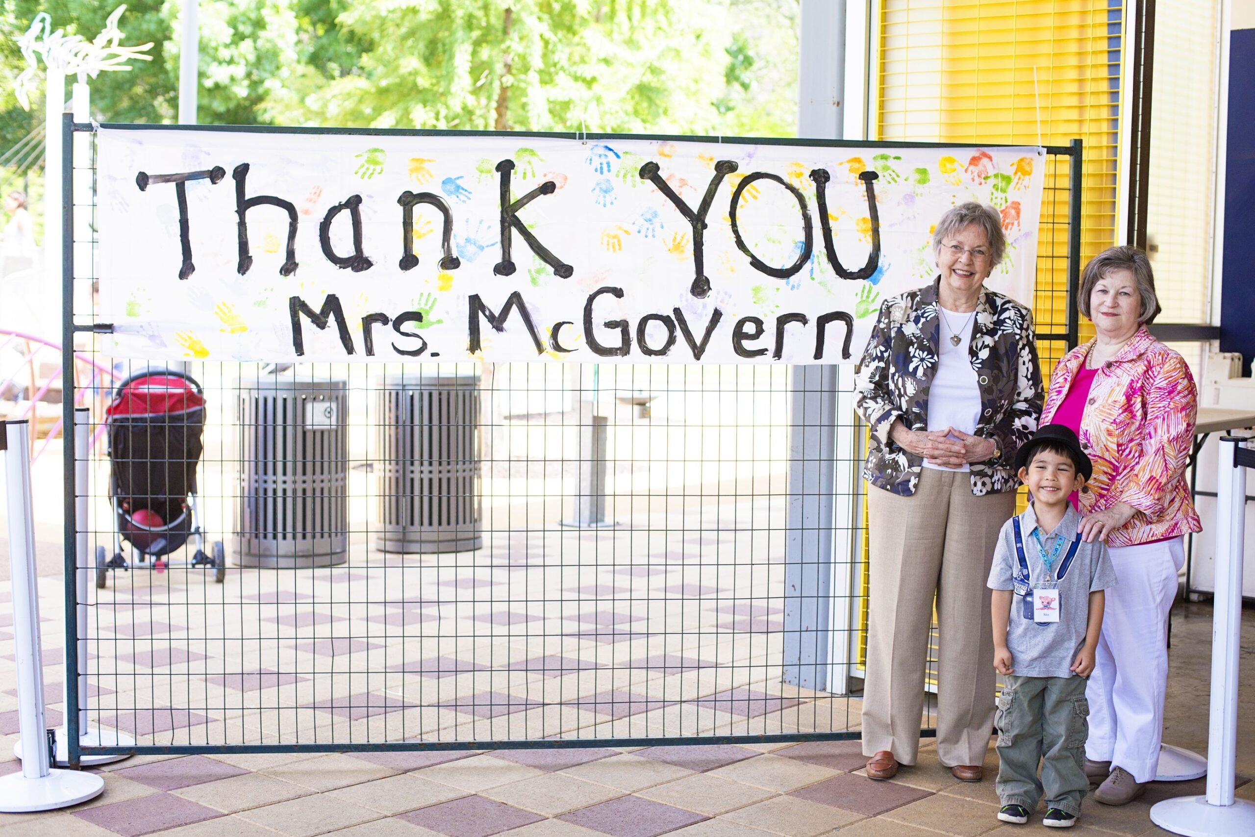 Kathrine McGovern posing with children in front of a poster reading "Thank you Mrs. McGovern" in front of the John P. McGovern playground at Discovery Green.